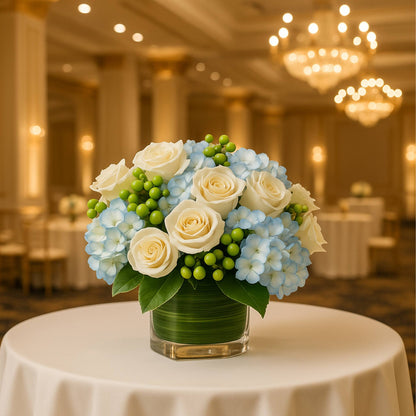 Floral arrangement with blue hydrangeas, white roses, and green berries on a table in a banquet hall.