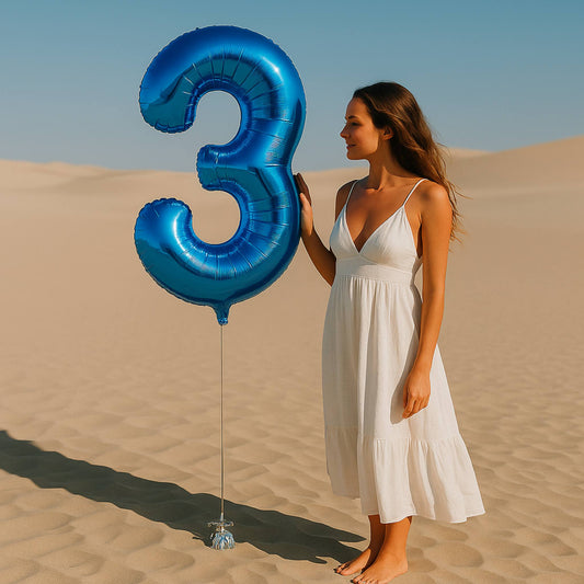 Woman in a white dress holding a large blue number 3 balloon in the desert.
