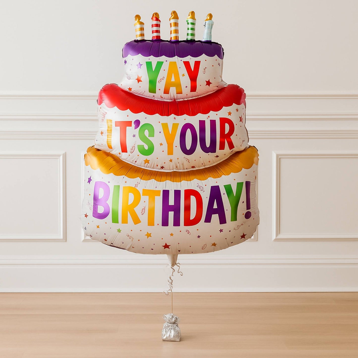 Colorful birthday cake-shaped balloon with candles against a white wall.