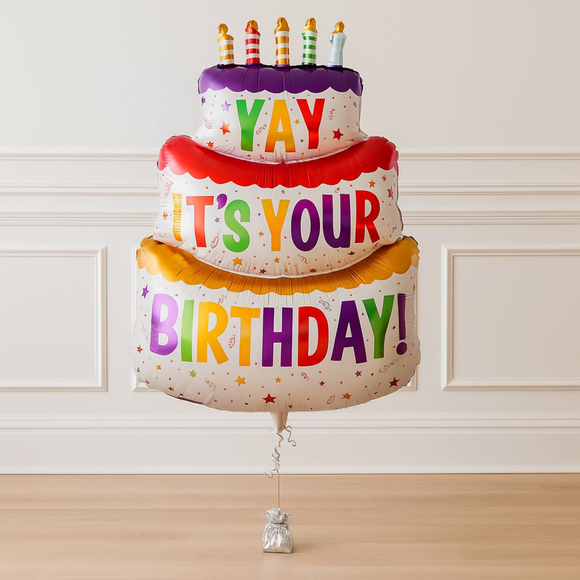 Colorful birthday cake-shaped balloon with candles against a white wall.