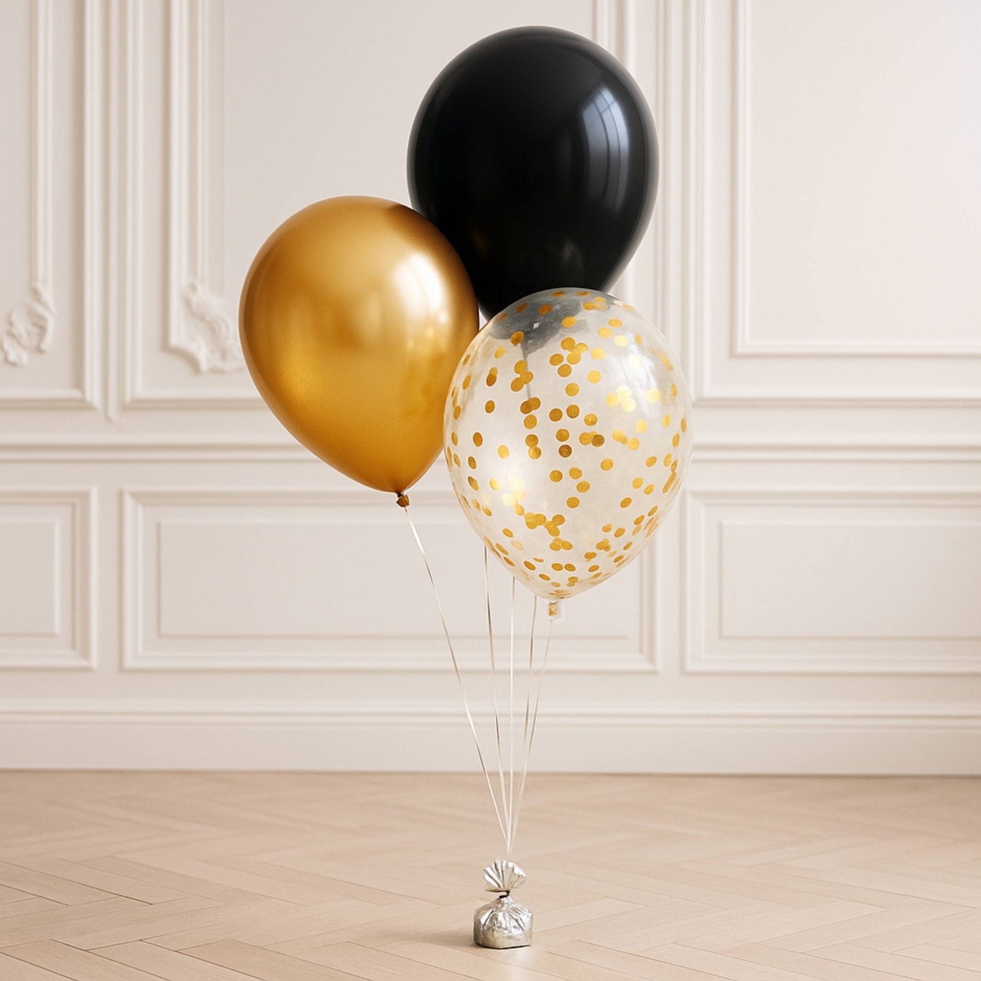 Three balloons in black, gold, and silver with confetti on a wooden floor against a white paneled wall.