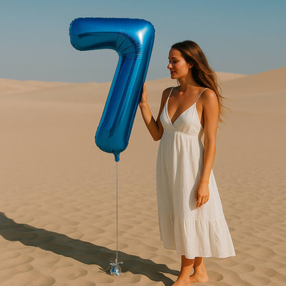 Woman in a white dress holding a large blue number 7 balloon in a desert setting