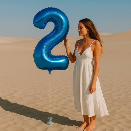 Woman in a white dress holding a large blue number two balloon in a desert setting