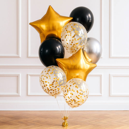 Decorative balloon bouquet with gold, black, and silver balloons on a wooden floor against a white paneled wall.