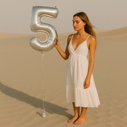 Woman in a white dress holding a large silver number 5 balloon in a desert setting