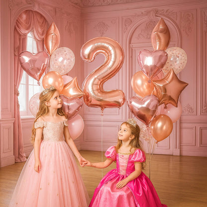Two young girls in pink dresses holding hands with rose gold balloons shaped like numbers and heart shapes in a decorated room.