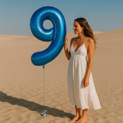 Woman in a white dress holding a large blue balloon shaped like the number 9 in a desert setting.