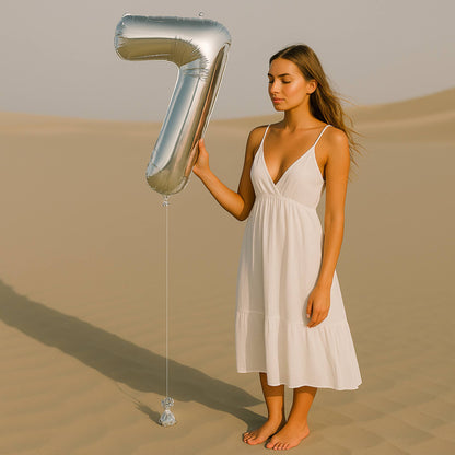 Woman in a white dress holding a large silver number 7 balloon in a desert setting