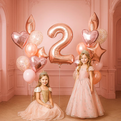 Two young girls in formal dresses standing in a room with pink balloons and a large number '2' balloon.