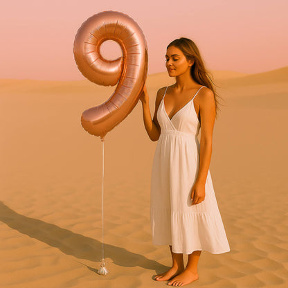 Woman in a white dress holding a large rose gold number 9 balloon in a desert setting