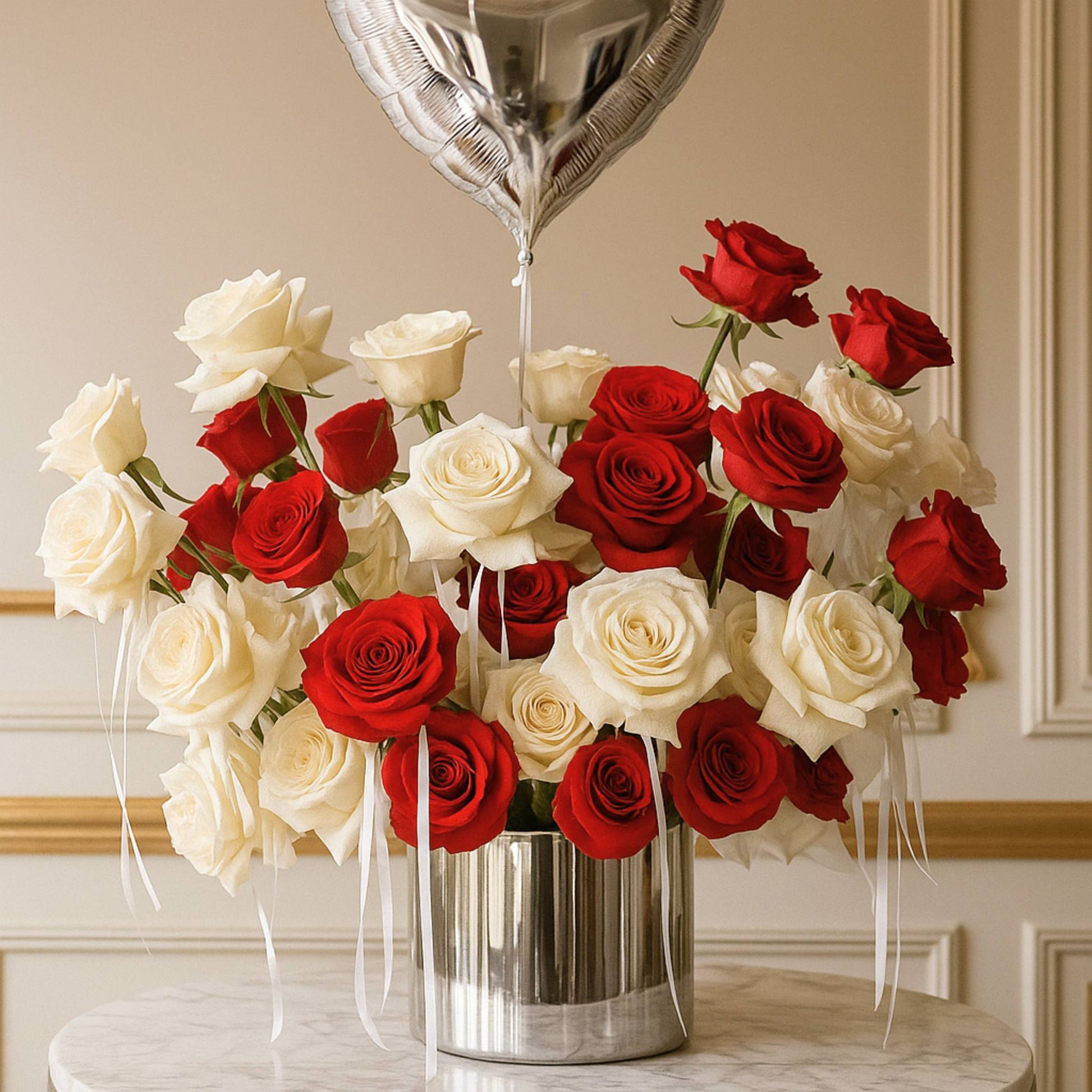 Bouquet of red and white roses in a silver vase with a silver heart-shaped balloon against a beige wall.