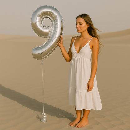 Woman in a white dress holding a large silver number '9' balloon in a desert setting