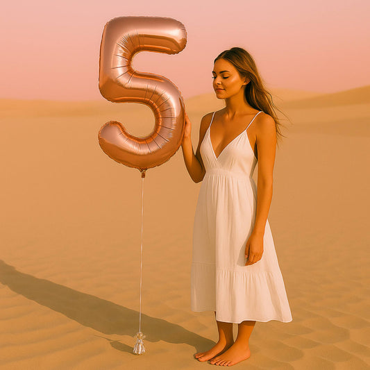Woman in a white dress holding a large rose gold number five balloon in a desert setting.