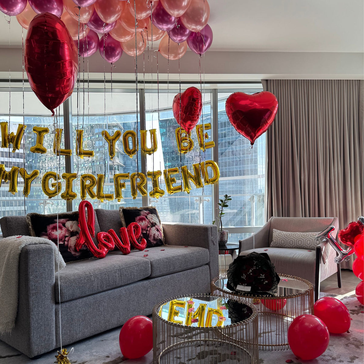 A room decorated with a variety of balloons, including heart-shaped balloons in red and pink, and balloons with 'Will you be my girlfriend' and 'love' text in golden color, creating a romantic atmosphere.