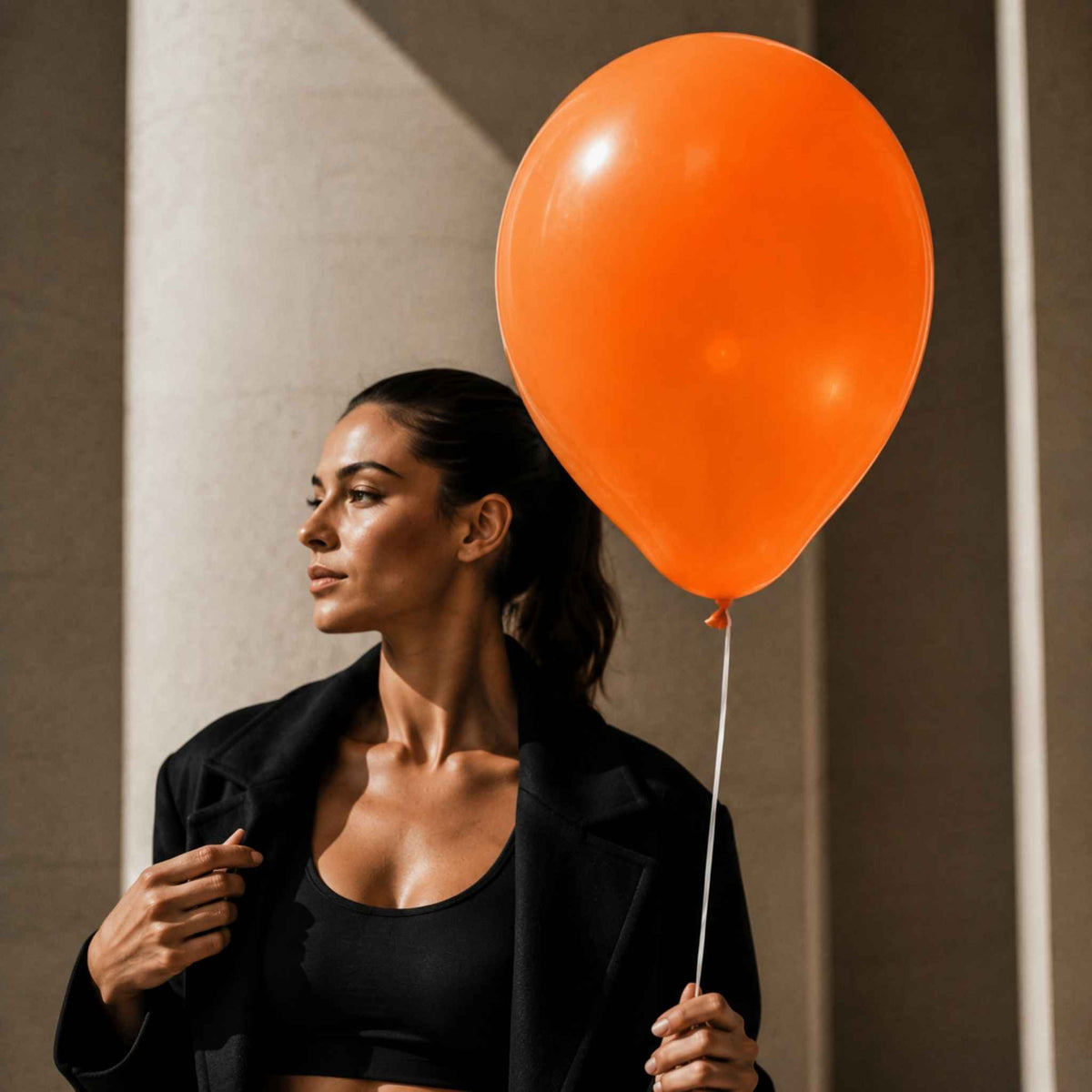 A woman in a black blazer and black sports bra holds a large, vibrant orange balloon next to her, standing against a neutral beige wall with dramatic shadows.