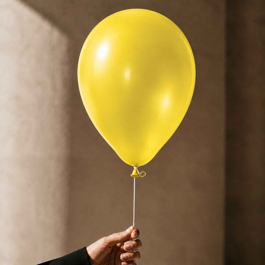 A single matte yellow balloon held by a hand against a warm beige wall with soft shadows.