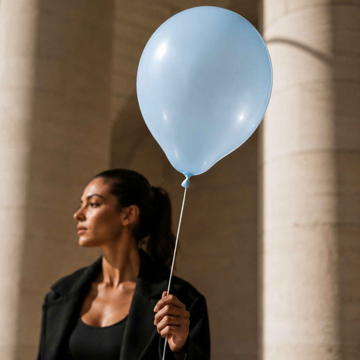 A woman in a black blazer and black top holds a light blue balloon against a background of warm beige stone columns.