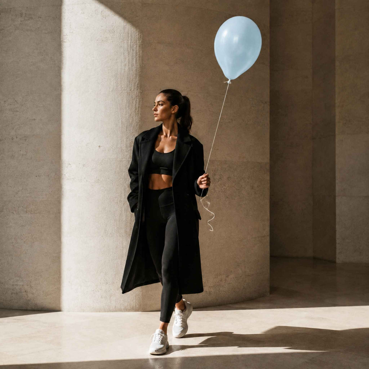 A woman in a black activewear set with a long black coat walks through a sunlit concrete corridor while holding a light blue balloon.