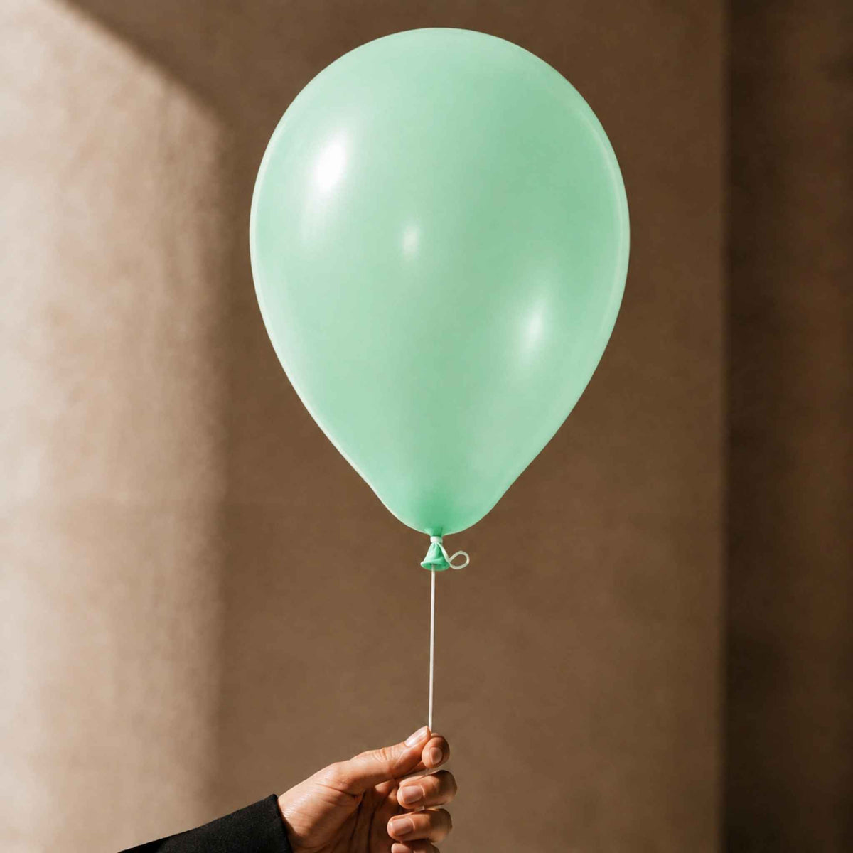 A mint green latex balloon held by a hand against a warm, textured brown background.