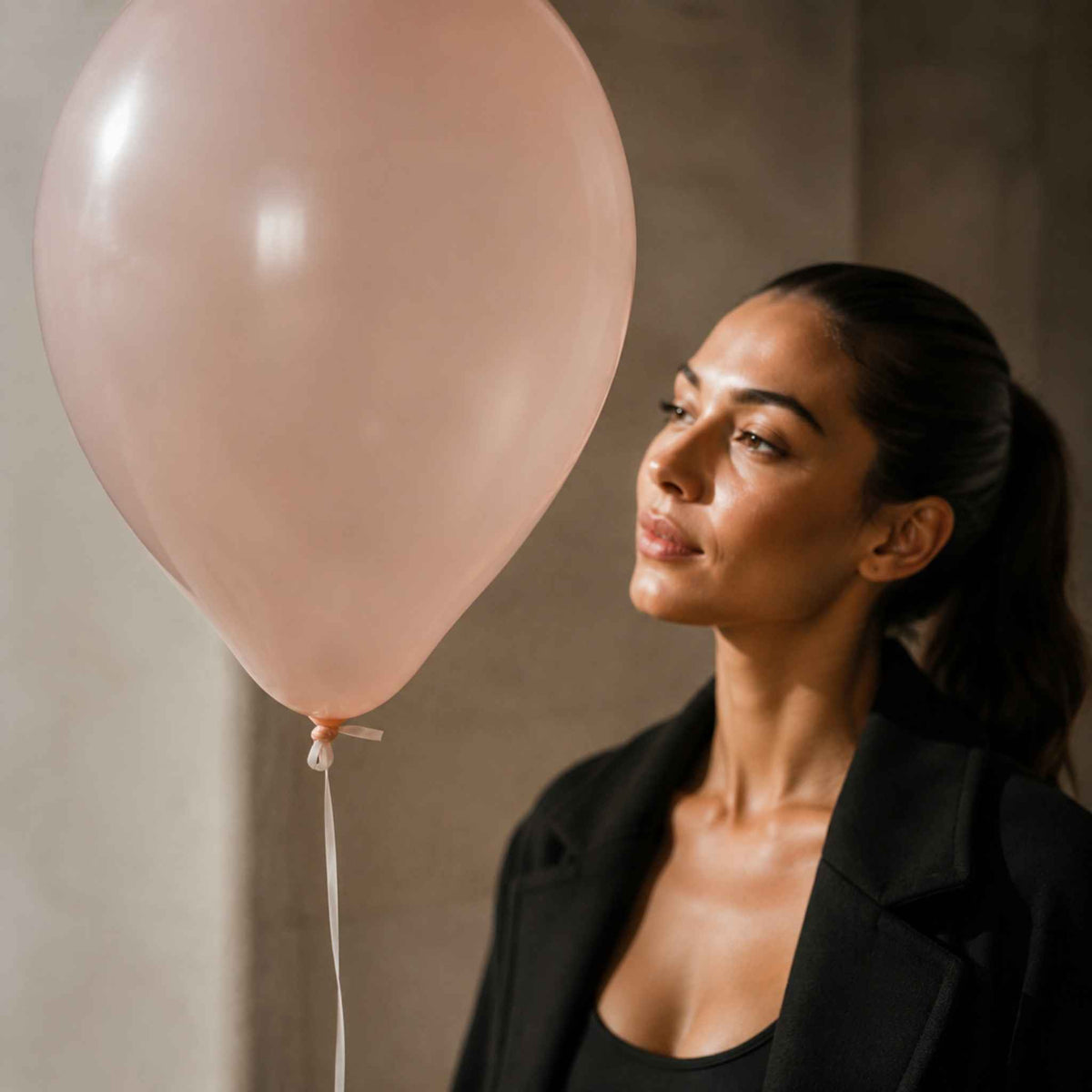 A woman with her hair in a ponytail wearing a black blazer and top, looking at a large pale pink balloon next to her against a neutral concrete wall.