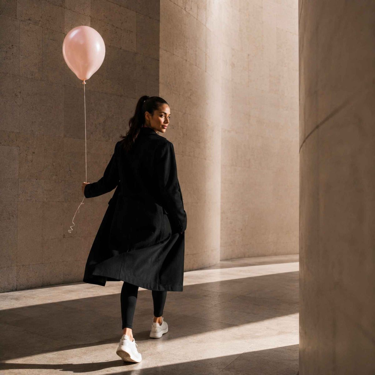 A woman in a black trench coat, black leggings, and white sneakers walks through a sunlit stone corridor while holding a light pink balloon.
