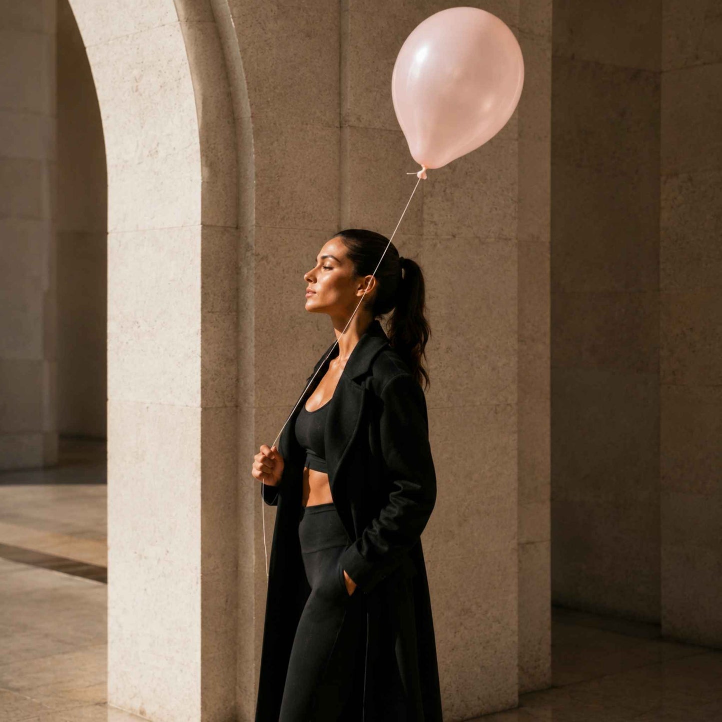 A woman in a black tailored outfit stands in a stone architectural corridor holding a pale pink balloon.