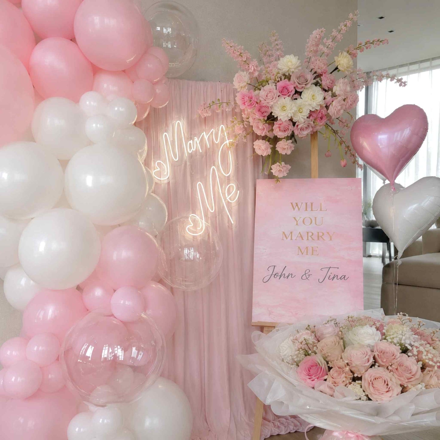 Decorative setup with pink and white balloons, flowers, and a 'Will You Marry Me' sign.