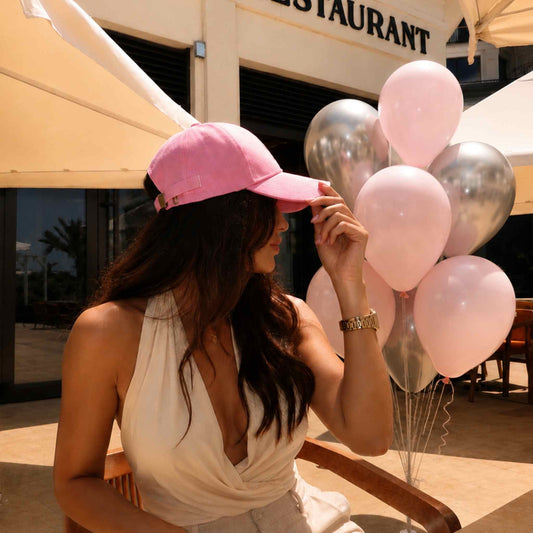 A woman wearing a pink baseball cap sits at an outdoor cafe next to a cluster of light pink and metallic silver balloons.