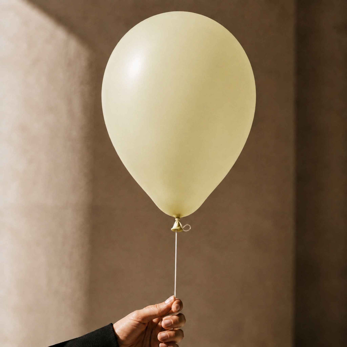 A pale cream-colored latex balloon held by a hand against a warm, textured brown background.