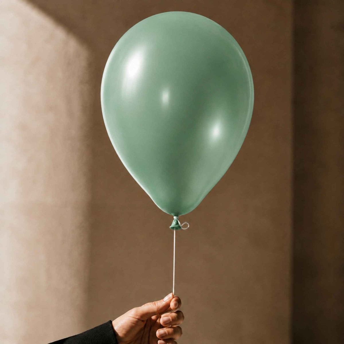 A matte sage green balloon held by a hand against a warm, textured beige wall.