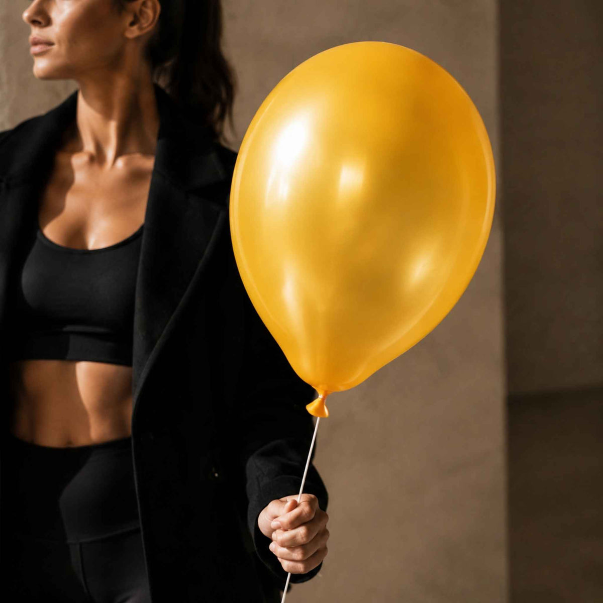 A woman in a black blazer and sports bra holds a large, shiny gold balloon against a warm, neutral-toned indoor background.