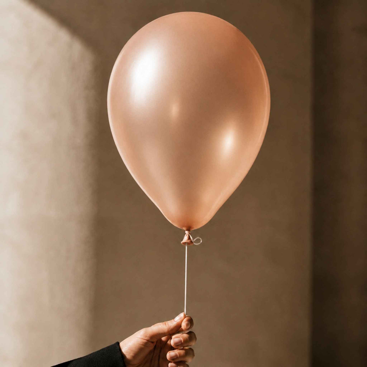 A single matte rose gold balloon held by a hand against a warm, textured beige wall.