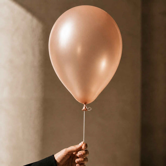 A single matte rose gold balloon held by a hand against a warm, textured beige wall.