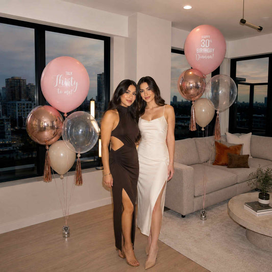 Two women posing in a modern living room decorated for a 30th birthday celebration, with pink and metallic rose gold balloon arrangements in the background.