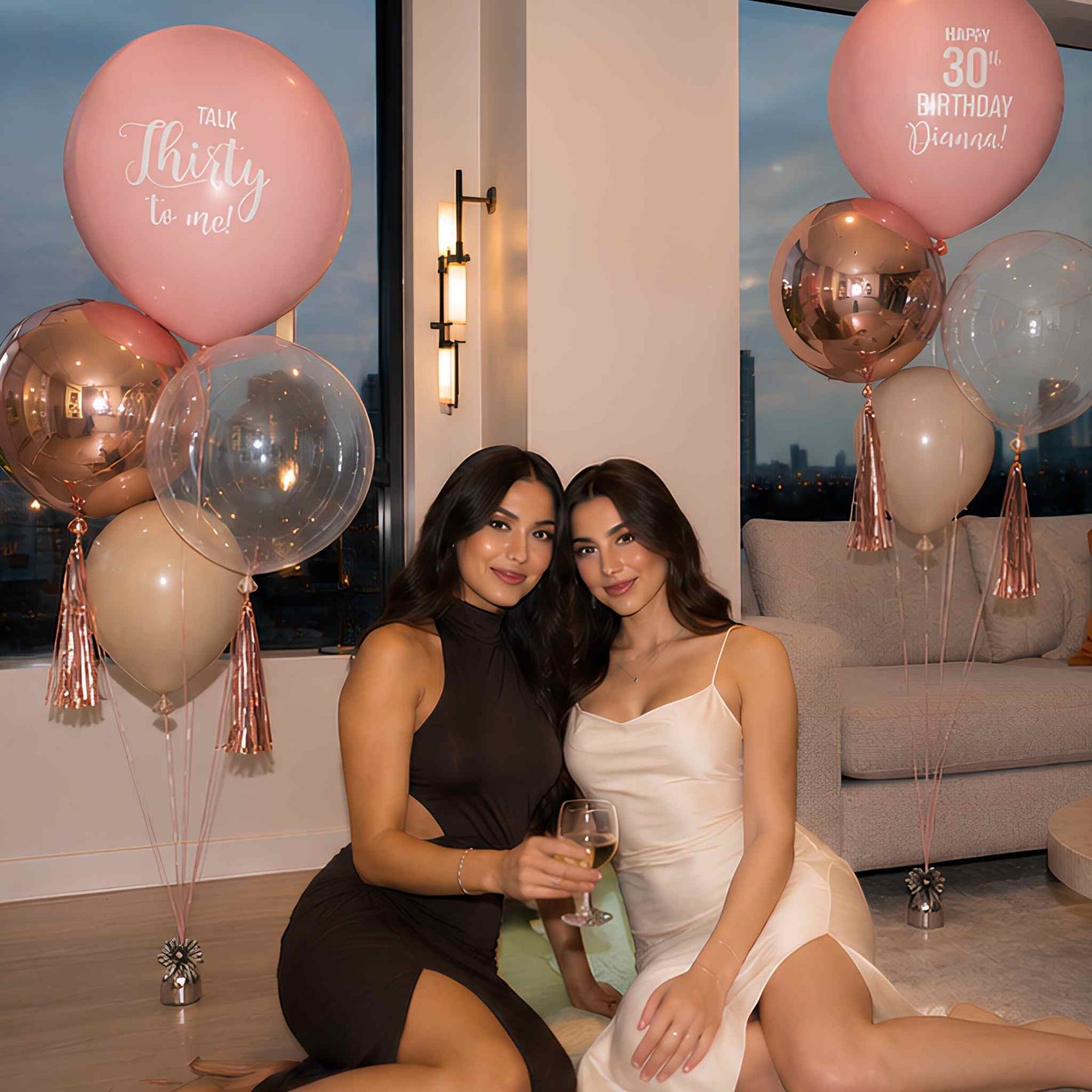 Two women posing indoors with birthday balloons. One woman wears a black dress and the other wears a white dress.