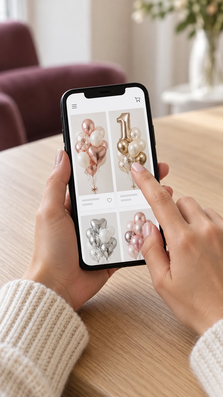 A person holds a smartphone over a wooden table, showing a screen with balloon decoration options in rose gold, gold, silver, and pink. The background shows a softly blurred indoor setting with a sofa and flowers.