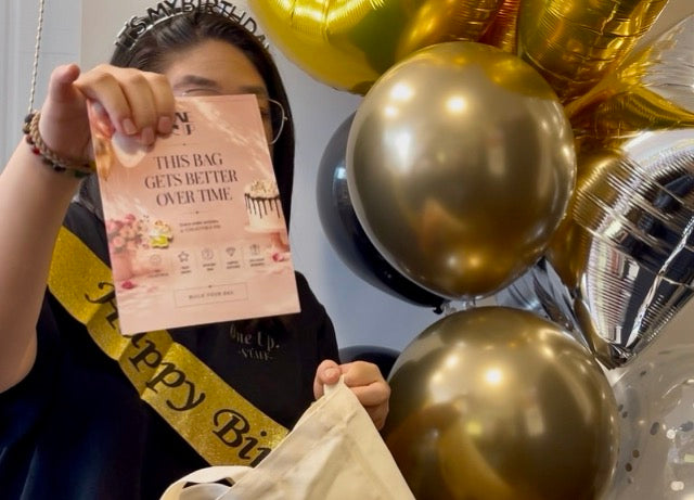 A person wearing a black t-shirt and a gold 'Happy Birthday' sash holds up a pink 'This Bag Gets Better Over Time' card next to a canvas tote bag, with gold and black balloons in the background.