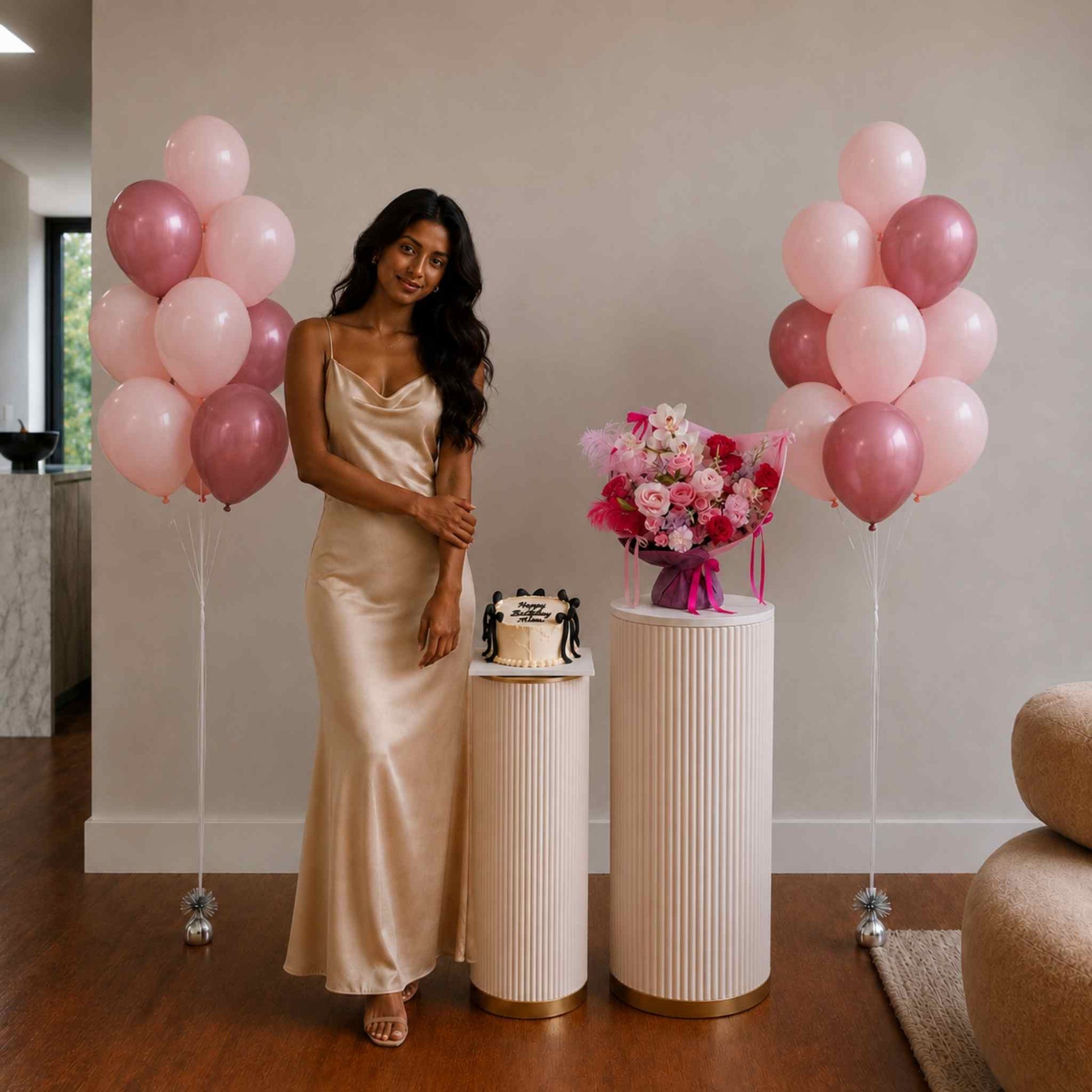 Woman in a gold dress standing between pink balloons with a cake and flowers on a table.