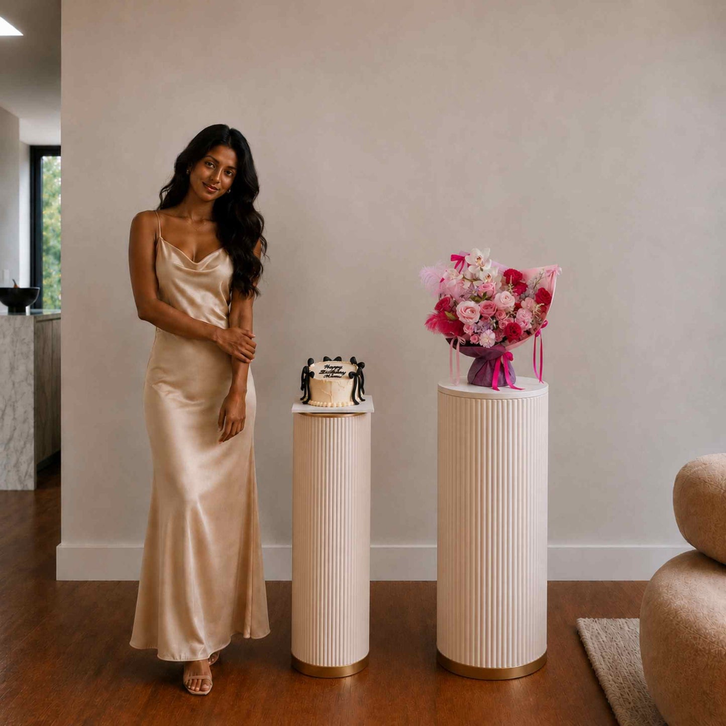 Woman in a long, light-colored dress standing next to a small cake and floral arrangement on a pedestal.