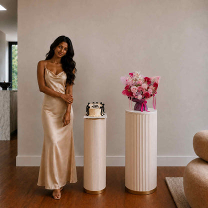 Woman in a long, light-colored dress standing next to a small cake and floral arrangement on a pedestal.