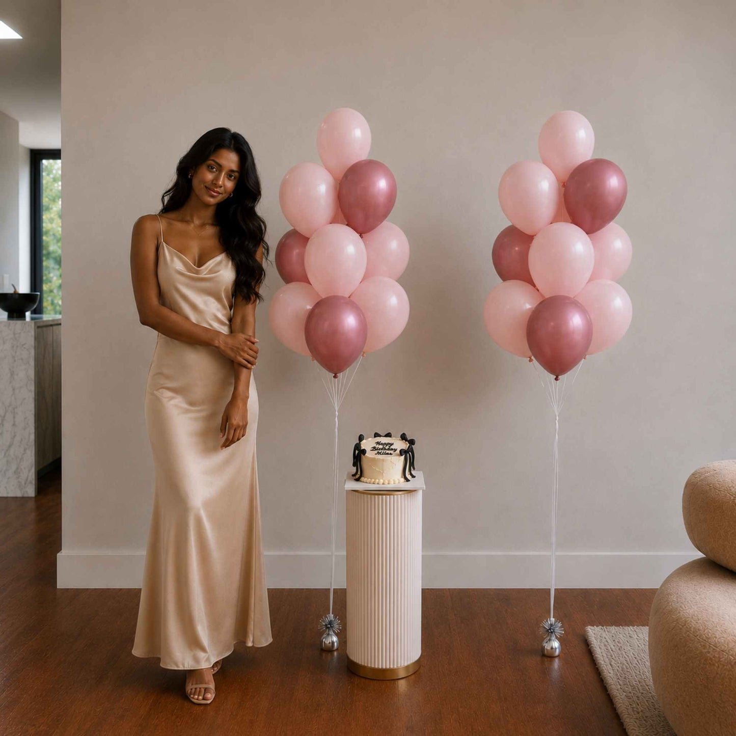 Woman in a long dress standing next to pink and gold balloons and a cake on a stand.