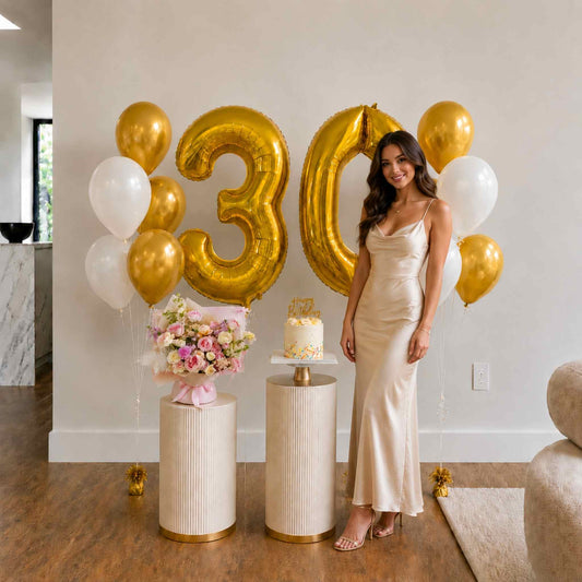 Woman in a gold dress standing next to '30' balloons and a cake in a room.