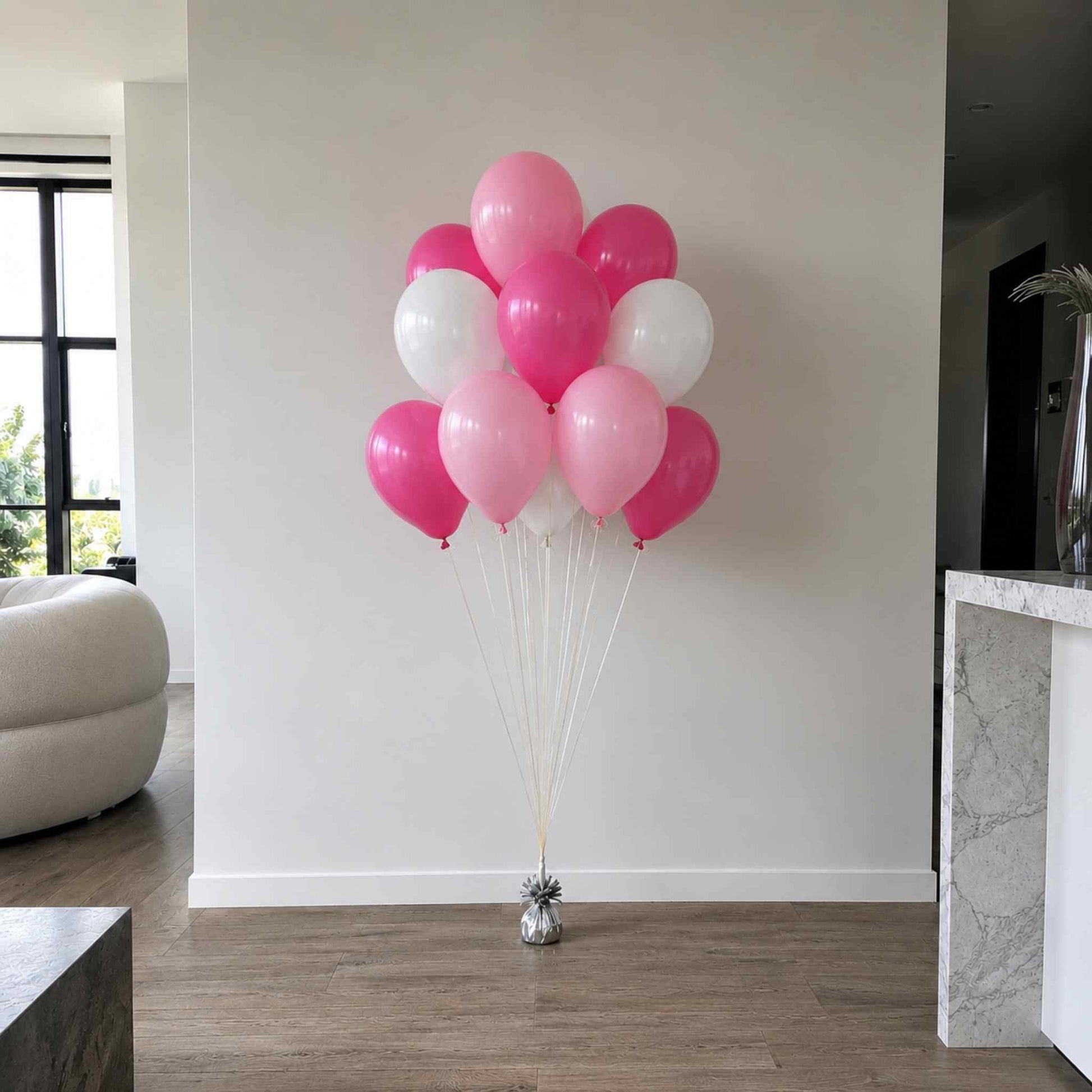 A cluster of pink and white balloons tied to a small metallic stand on a wooden floor in a modern room with a white wall and light-colored furniture.