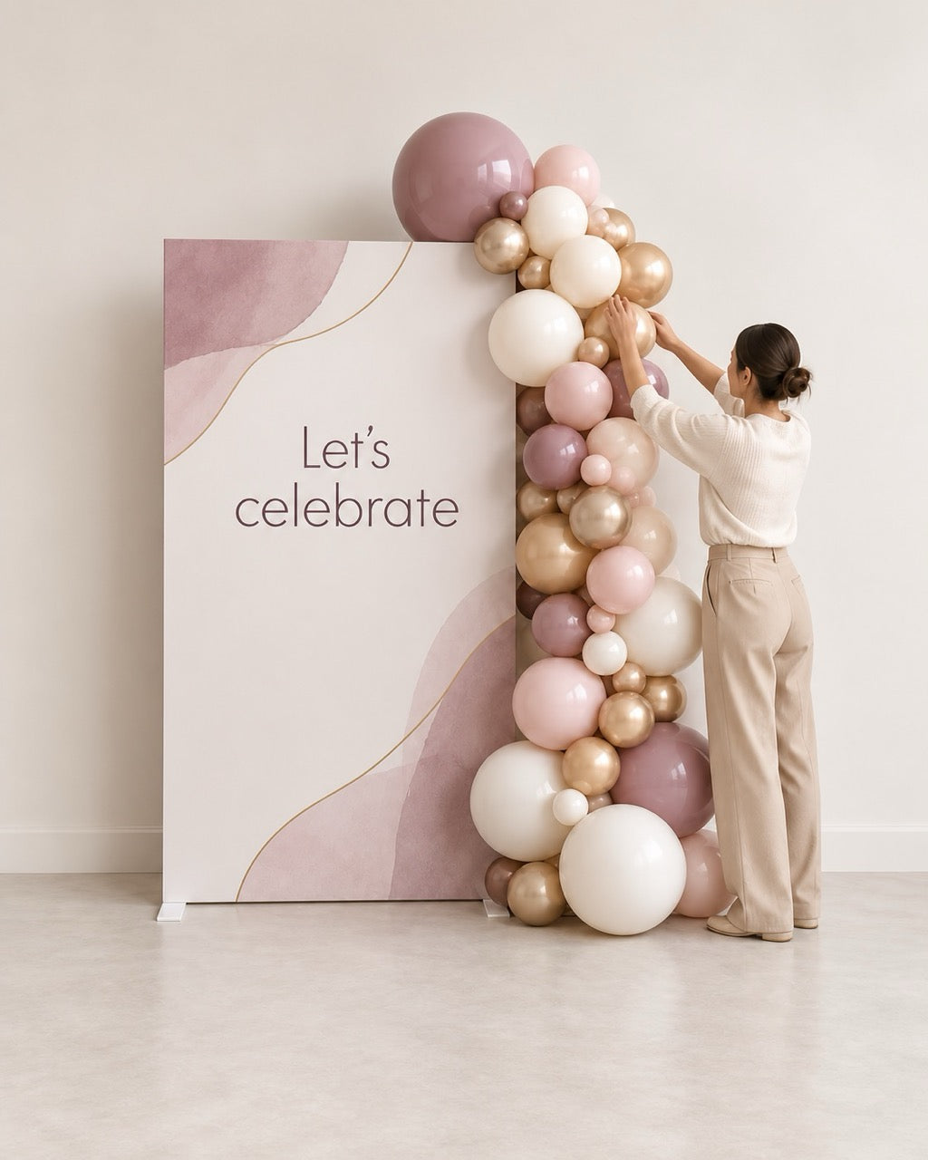 A woman in a white top and beige pants is adjusting a large balloon garland next to a printed backdrop panel that reads 'Let's celebrate'. The balloons are in soft blush pink, white, and metallic gold tones, arranged in a decorative arch against a clean, light beige wall and floor.
