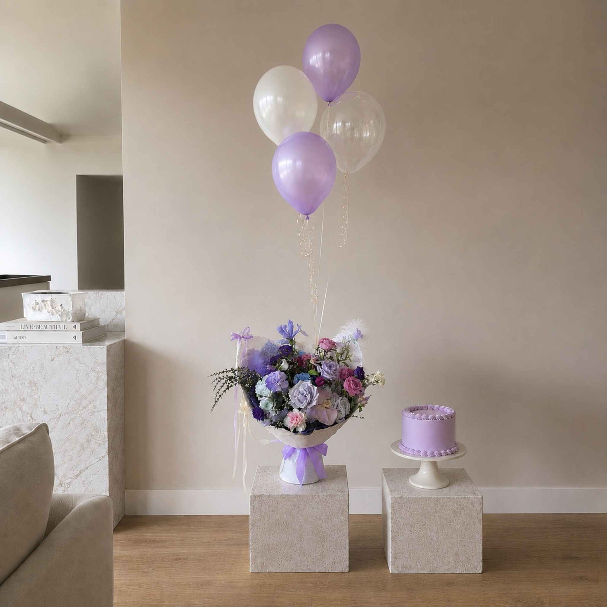 Bouquet of flowers and purple balloons on a stand with a small cake in a room.