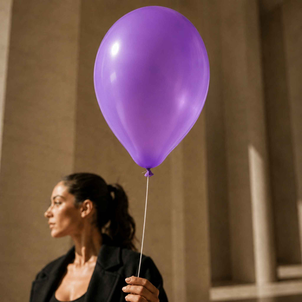 A woman in a black blazer holds a large, glossy purple balloon against a blurred beige architectural background.