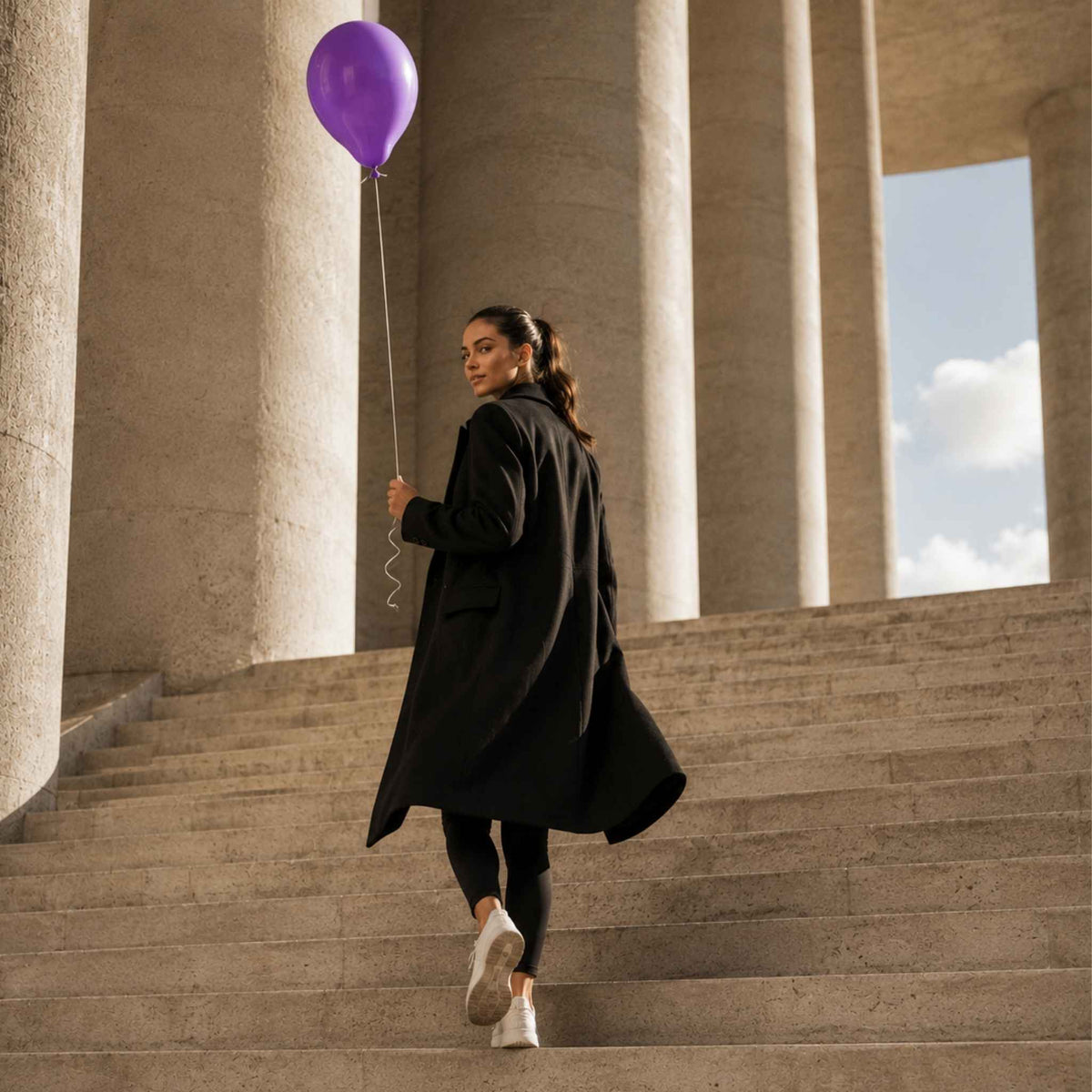 A woman in a long black coat walks down large stone steps while holding a purple balloon, with tall columns and an open sky in the background.