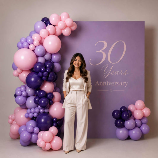 A woman stands in front of a large lavender backdrop reading '30 Years Anniversary,' decorated with pink and purple balloon garlands on both sides. She is wearing a light beige tailored jumpsuit or matching set.