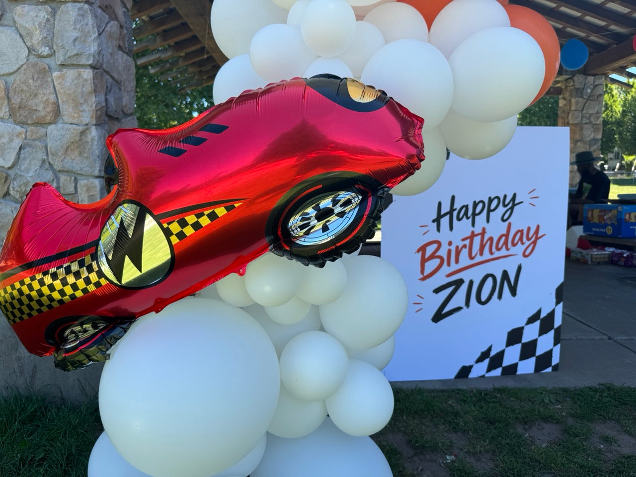 A red race car foil balloon with yellow and black checkered accents is displayed outdoors among white balloons, next to a white 'Happy Birthday Zion' sign.