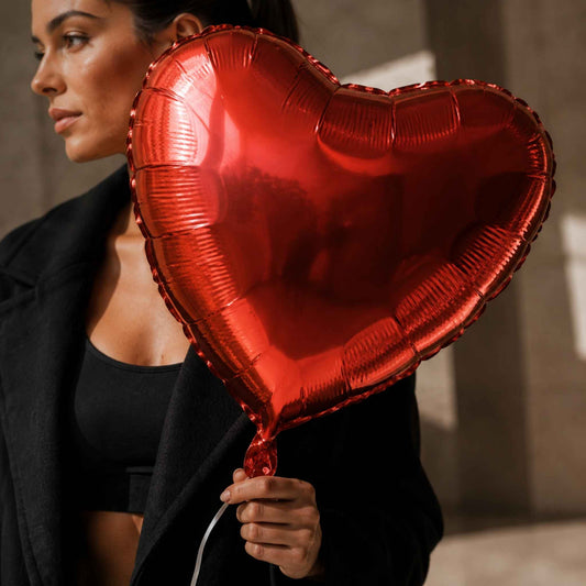 A woman in a black blazer and black top holds a large metallic red heart-shaped foil balloon, photographed in a softly lit indoor setting.